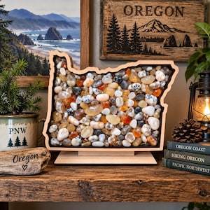 May include: A wooden display frame shaped like the state of Oregon, filled with a collection of smooth, polished river stones in shades of white, tan, orange, and grey. The frame sits on a flat wooden base, showcasing a variety of natural rock textures.