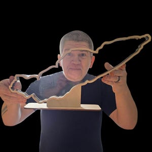 May include: A person holds a wooden cutout of a race track. The cutout is brown and has a black outline. The person is wearing a dark shirt.
