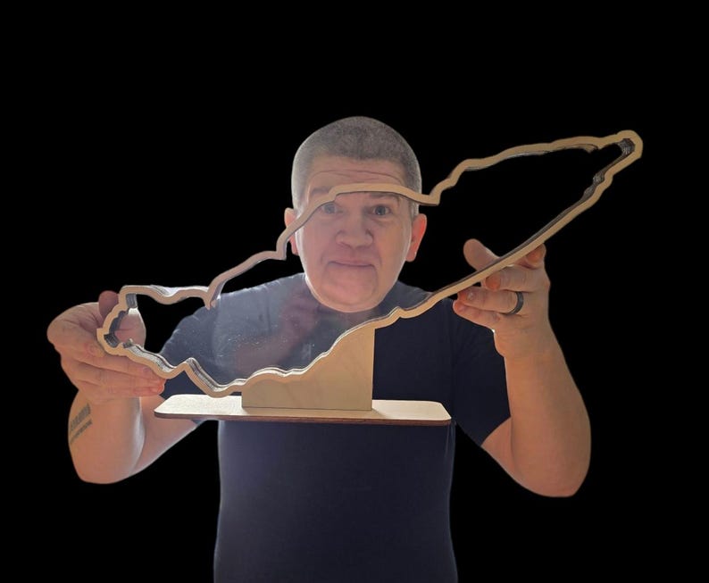 May include: A person holds a wooden cutout of a race track. The cutout is brown and has a black outline. The person is wearing a dark shirt.