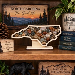 May include: A wooden cutout of the state of North Carolina filled with various polished stones, mounted on a rectangular base. The stones are a mix of brown, gray, white, and green hues, creating a mosaic effect within the state border outline.