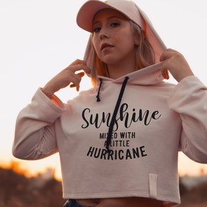 Puede incluir: Una sudadera con capucha rosa recortada con un cord&oacute;n de ajuste negro y el texto "Sunshine mixed with a little hurricane" impreso en la parte delantera.