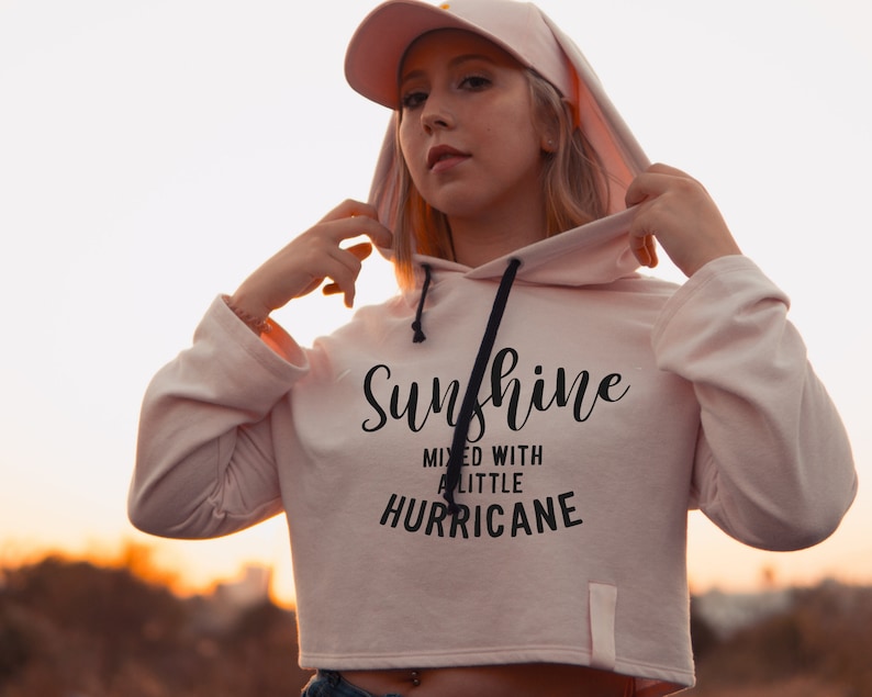 Puede incluir: Una sudadera con capucha rosa recortada con un cord&oacute;n de ajuste negro y el texto "Sunshine mixed with a little hurricane" impreso en la parte delantera.