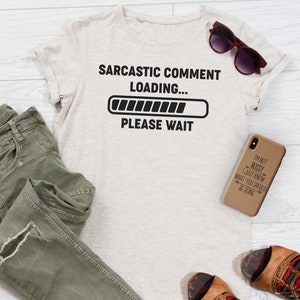 Puede incluir: Una camiseta beige con un gr&aacute;fico negro que dice "Comentario sarc&aacute;stico cargando..." con una barra de progreso y "Por favor, espere". Tambi&eacute;n se ve un par de jeans, una funda de tel&eacute;fono con el texto "No soy mandona, solo s&eacute; lo que deber&iacute;as estar haciendo", y unas sandalias.