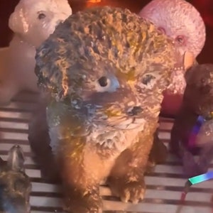 May include: A small, brown and white ceramic figurine of a dog with curly fur. The dog is sitting with its paws tucked under its body. The figurine is on a striped surface.