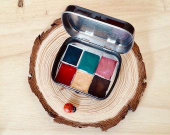 May include: A small, open, silver metal tin containing six squares of colorful paint. The colors include dark blue, green, red, gold, brown, and pink. The tin sits on a wooden slice with a small ladybug figurine.