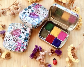May include: Three floral-patterned metal watercolor paint tins. One tin is open, revealing six vibrant paint colors: green, yellow, pink, red, and blue. The other two tins are closed. Dried flowers and a ladybug figurine are scattered around the tins.