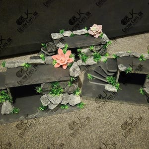 May include: A three-tiered slate rock reptile habitat with a cave opening on each level. The habitat is decorated with artificial plants and pink flowers.