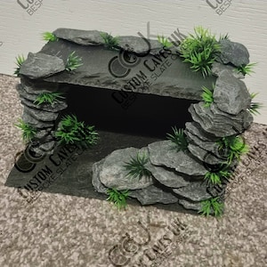 May include: A grey slate rock cave background for a reptile terrarium. The cave is made of stacked slate rocks and has a small opening. The cave is surrounded by green plastic plants.