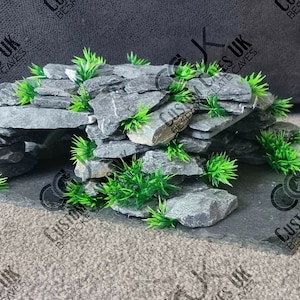 May include: A grey slate rock cave background for a terrarium or reptile enclosure. The cave is made of stacked slate rocks with a small opening at the front. The cave is decorated with green plastic plants.