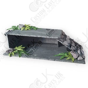 May include: A grey slate reptile habitat with a two-tiered platform and a cave opening. The habitat features a small rock wall and green plastic plants.