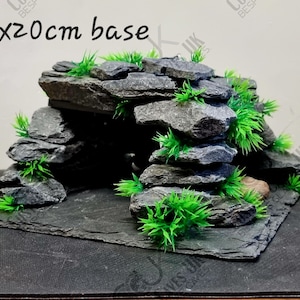 May include: A grey slate rock cave background for a terrarium or reptile enclosure. The cave is 20cm by 20cm and features a realistic rock formation with green plastic plants.
