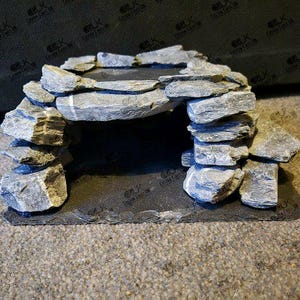 May include: A grey stone archway structure, possibly for aquarium or terrarium decoration. The arch is made from stacked, rough-hewn stones, giving a natural look. The base is a dark, rectangular slate. The design is intended to resemble a miniature cave.
