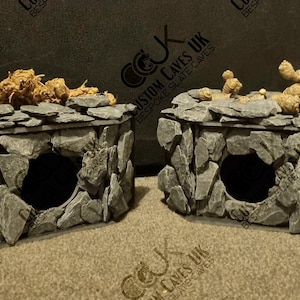 May include: Two grey stone reptile hides with a rounded opening. The hides are made of stacked slate stones and have a rough texture. The hides are placed on a brown surface.