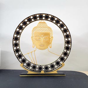 Könnte beinhalten: Eine goldfarbene Buddha-Kopf-Skulptur in einem schwarzen kreisförmigen Rahmen mit sternförmigen Ausschnitten. Die Skulptur ist auf einem schwarz-goldenen Sockel montiert und schafft ein dekoratives und spirituelles Stück.