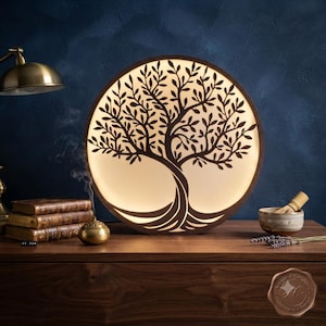 Peut inclure: Une lampe circulaire en bois présentant la silhouette rétroéclairée d'un arbre aux branches et racines complexes. La lampe émet une lueur chaude, soulignant le cadre en bois sombre. Elle est posée sur une surface en bois avec des livres et un bol.