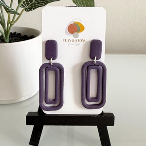 May include: A pair of dark purple rectangular earrings with a double-frame design. The earrings are displayed on a white card with the text "CLAY & STONE" and a logo. The earrings are showcased on a small black stand.