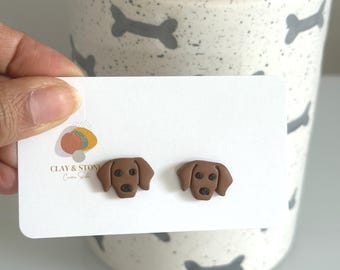 May include: A pair of brown dog-shaped stud earrings displayed on a white rectangular card. The earrings feature a smooth finish with black details for the eyes and nose. The card includes a logo with the text CLAY & STONE Creative Studio.