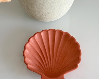 Puede incluir: Un plato en forma de concha de color coral con una superficie texturizada. El plato tiene un borde festoneado y un patrón en forma de abanico que irradia desde el centro. El plato es probablemente de cerámica o un material similar.