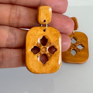 May include: A pair of gold ceramic earrings with a geometric design. The earrings are rectangular with a cutout in the center that resembles a flower.