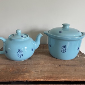 May include: A light blue ceramic teapot and a small lidded pot with a matching design. Both pieces have a tulip pattern in dark blue. The teapot has a curved spout and handle. The pot has two small handles.