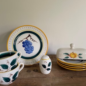 May include: A set of vintage ceramic kitchenware featuring a grape motif. The set includes a large plate, two cups and saucers, a salt shaker, and a butter dish with a lid. The pieces are decorated with hand-painted grapes, leaves, and vines in shades of green, blue, and yellow.