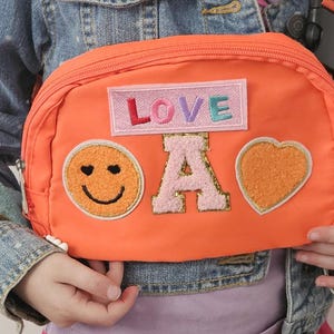 May include: Orange fanny pack with a smiley face, the letter A, a heart, and a pink patch that says "LOVE" on it.