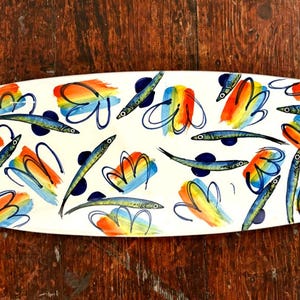 Puede incluir: Plato rectangular de cerámica con fondo blanco. El plato está decorado con diseños florales abstractos azules, naranjas y amarillos y peces azules. El plato tiene esquinas redondeadas y está colocado sobre una superficie de madera.