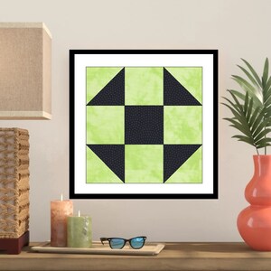 May include: Framed artwork featuring a geometric quilt block design. The quilt block has a central black square surrounded by green squares and black triangles. The artwork is displayed on a wooden surface with candles, glasses, and a vase.