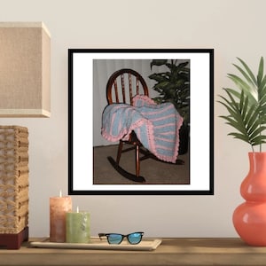 May include: A handmade, striped crochet blanket in light blue and pink rests on a wooden rocking chair. The blanket has a scalloped edge and is draped over the chair's seat and back. The chair is set against a neutral background.