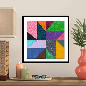 May include: A framed quilt block with a geometric design. The quilt features various fabric patterns and colors, including pink, blue, green, and yellow. The artwork is displayed on a wooden surface with candles, glasses, and a lamp.