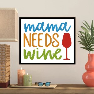 May include: A framed cross-stitch artwork with the words "mama NEEDS wine" in blue, orange, and green. A red wine glass graphic is to the right. The artwork is displayed on a wooden surface with candles, glasses, and a vase.