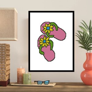 May include: A framed artwork featuring a pixelated design of two pink flip-flops, each adorned with a yellow and blue flower. The flip-flops have green straps. The artwork is displayed on a wooden surface with candles and a lamp.