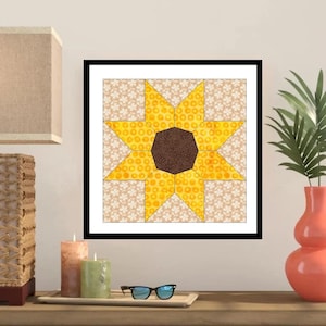Könnte beinhalten: Ein gerahmter Sonnenblumen-Quiltblock mit schwarzem Rand. Die Sonnenblume hat eine braune Mitte und gelbe Blütenblätter mit einem gepunkteten Muster. Der Hintergrund ist ein beiger Stoff mit weißem Blumenmuster.