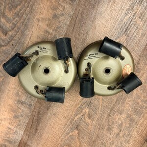 May include: Two circular, gold-toned metal ceiling light fixture bases, each featuring three black plastic light bulb sockets connected by short wires. Each base has a central hole and text printed on the surface stating 60 WATTS PER SOCKET.