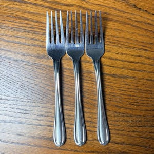 May include: Three silver-colored metal forks arranged side by side. Each fork features four tines and a smooth, slender handle with a subtle decorative detail at the base. The polished finish reflects light, showing a clean and simple design.