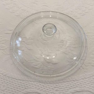 Puede incluir: Una tapa circular de vidrio transparente con una superficie lisa ligeramente abovedada y un pomo central elevado. La tapa presenta un borde exterior distintivo diseñado para encajar de forma segura en un recipiente, con un acabado traslúcido.
