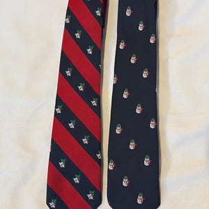 May include: Two neckties displayed side by side. The left tie features diagonal red and navy blue stripes with a repeating pattern of white bells and green holly. The right tie is solid navy blue with a repeating pattern of small snowmen.