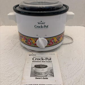 May include: A white Rival Crock-Pot stoneware slow cooker with a clear glass lid, side handles, and a front control knob. The base features a colorful chili pepper pattern. An accompanying owner's guide manual is placed in front of the appliance.