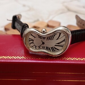 May include: A silver-toned wristwatch with a distorted oval face and black Roman numerals. The watch has a black leather band and the text "DeBurgh" is visible on the watch face. The watch is resting on a red box.