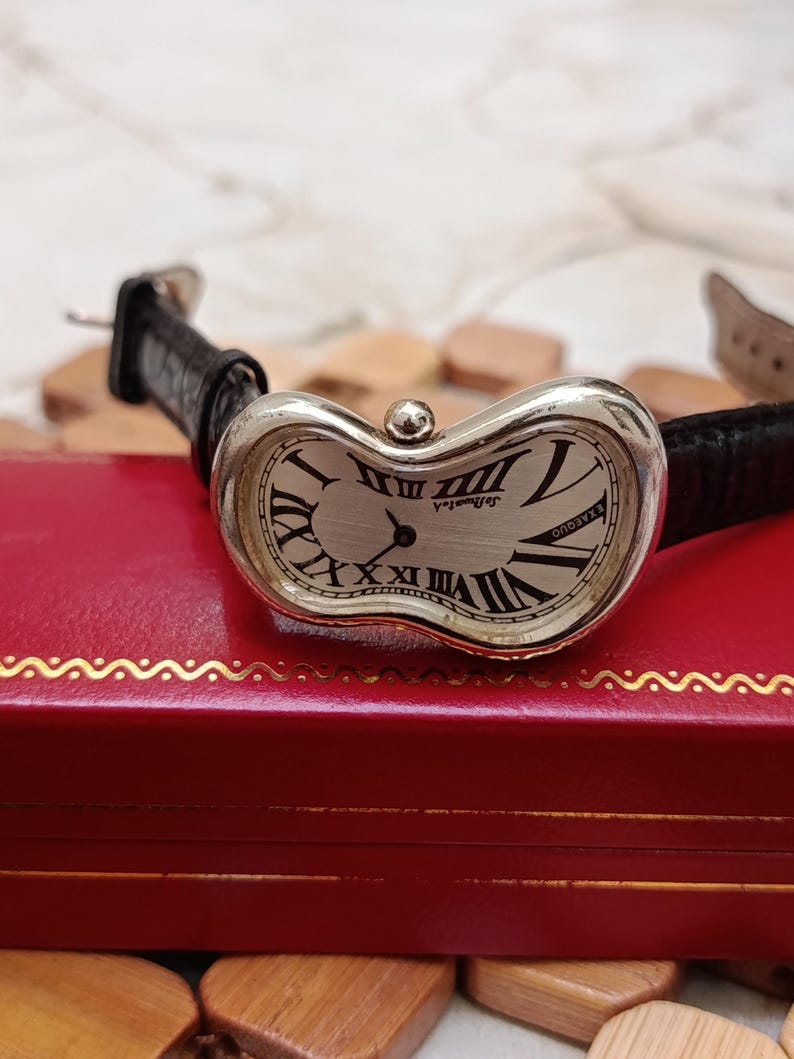 May include: A silver-toned wristwatch with a distorted oval face and black Roman numerals. The watch has a black leather band and the text "DeBurgh" is visible on the watch face. The watch is resting on a red box.