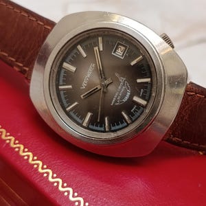 May include: A silver-toned wristwatch with a brown leather strap and a dark gray dial. The watch face features the text 'VERYWATCH', 'MINERAL CRYSTAL', '30 ATMOS', 'SQUALE', and 'SWISS MADE', with a date window at the three o'clock position.