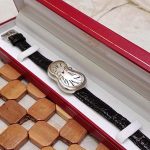 May include: A silver-toned wristwatch with a black leather band, presented in a red box. The watch face has Roman numerals and a unique, curved design. The watch band is approximately 20 cm long.