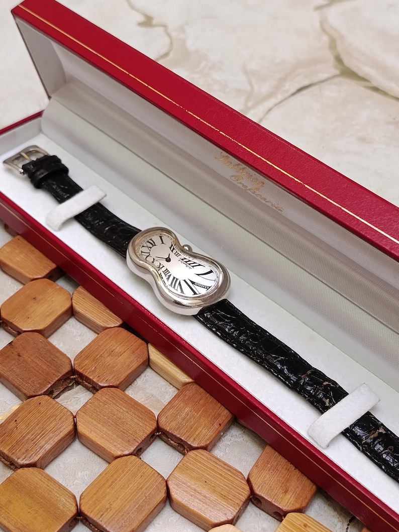 May include: A silver-toned wristwatch with a black leather band, presented in a red box. The watch face has Roman numerals and a unique, curved design. The watch band is approximately 20 cm long.
