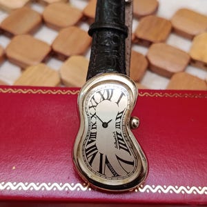 May include: A silver-toned wristwatch with a melting, curved face and black Roman numerals. The watch has a black leather band and the words "Softwatch" and "Exaequo" are visible on the face. The watch is resting on a red box.