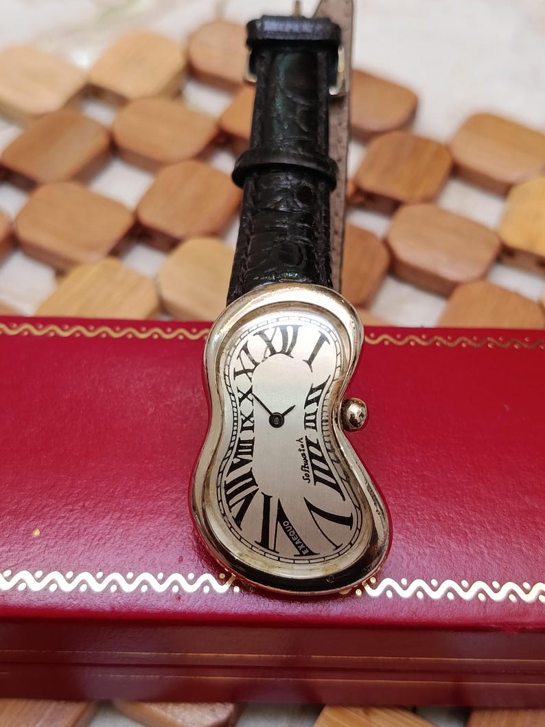 May include: A silver-toned wristwatch with a melting, curved face and black Roman numerals. The watch has a black leather band and the words "Softwatch" and "Exaequo" are visible on the face. The watch is resting on a red box.