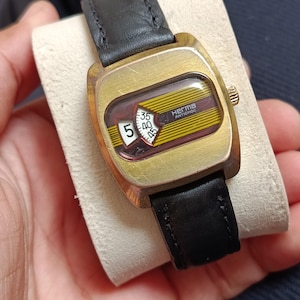May include: A vintage Herma Antichoc wristwatch with a gold-tone cushion-shaped case and a black leather strap. The dial features a horizontal yellow and brown striped pattern with a white window displaying the number 5 and a minute indicator.