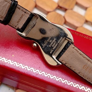 May include: Close-up of a watch with a silver-tone buckle and a black leather strap. The watch's back is visible, with the text "Softwatch" and other markings. The watch is resting on a red surface.