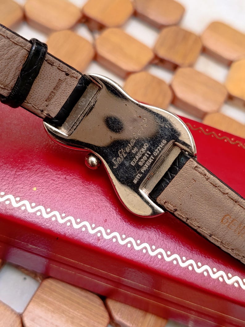 May include: Close-up of a watch with a silver-tone buckle and a black leather strap. The watch's back is visible, with the text "Softwatch" and other markings. The watch is resting on a red surface.