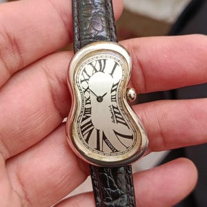 May include: A silver-toned wristwatch with a unique, melting design. The watch face features Roman numerals and the text "EXAEQUO" and "Softwatch". The black leather band has a textured, embossed pattern.