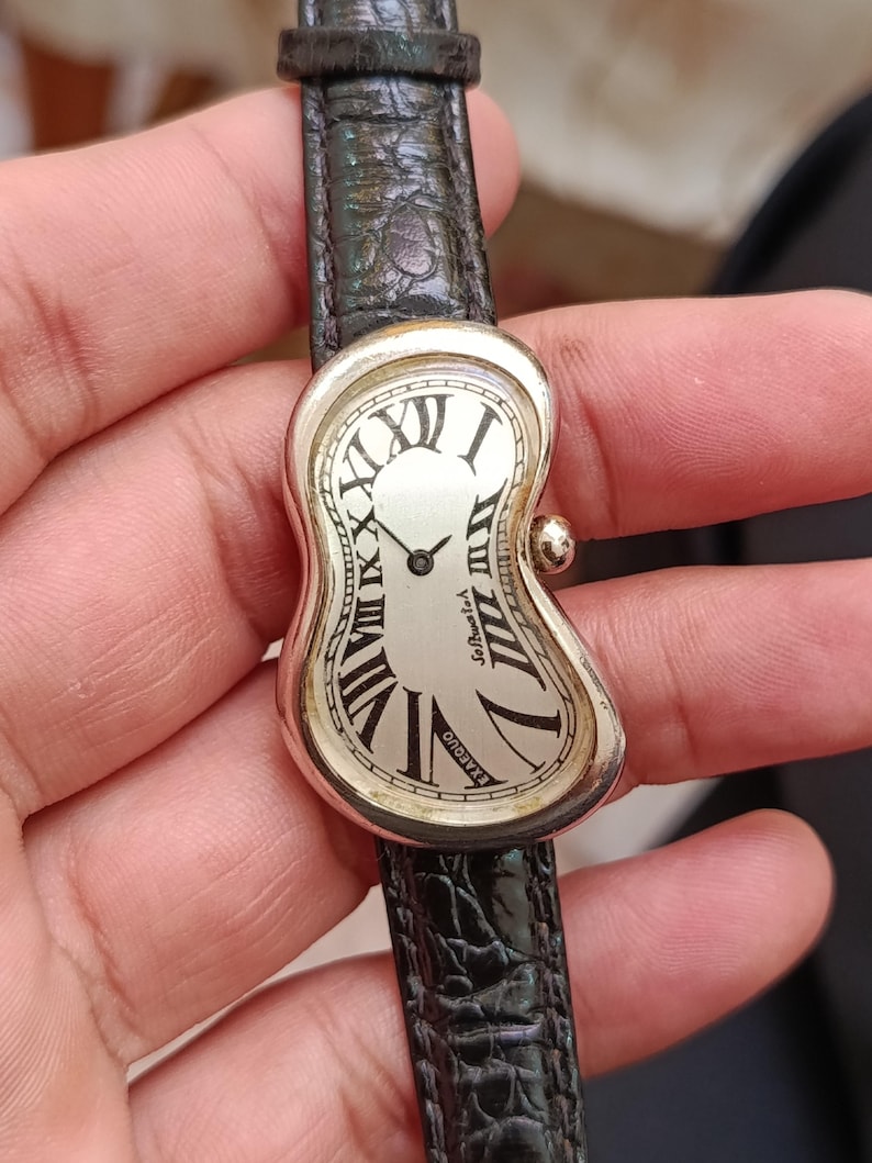 May include: A silver-toned wristwatch with a unique, melting design. The watch face features Roman numerals and the text "EXAEQUO" and "Softwatch". The black leather band has a textured, embossed pattern.